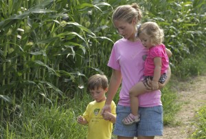 Mary holding walking with her two oldest children by a corn field.